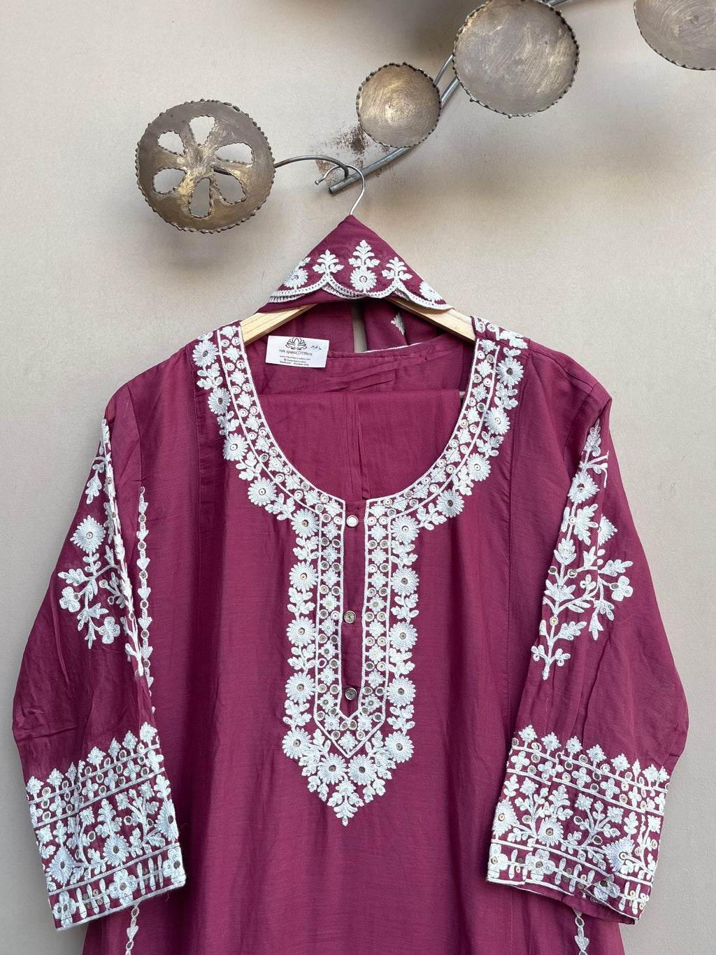 A deep magenta-purple cotton kurta with intricate white floral embroidery on the neckline, sleeves, and cuffs, hanging on a wooden hanger against a textured beige wall.