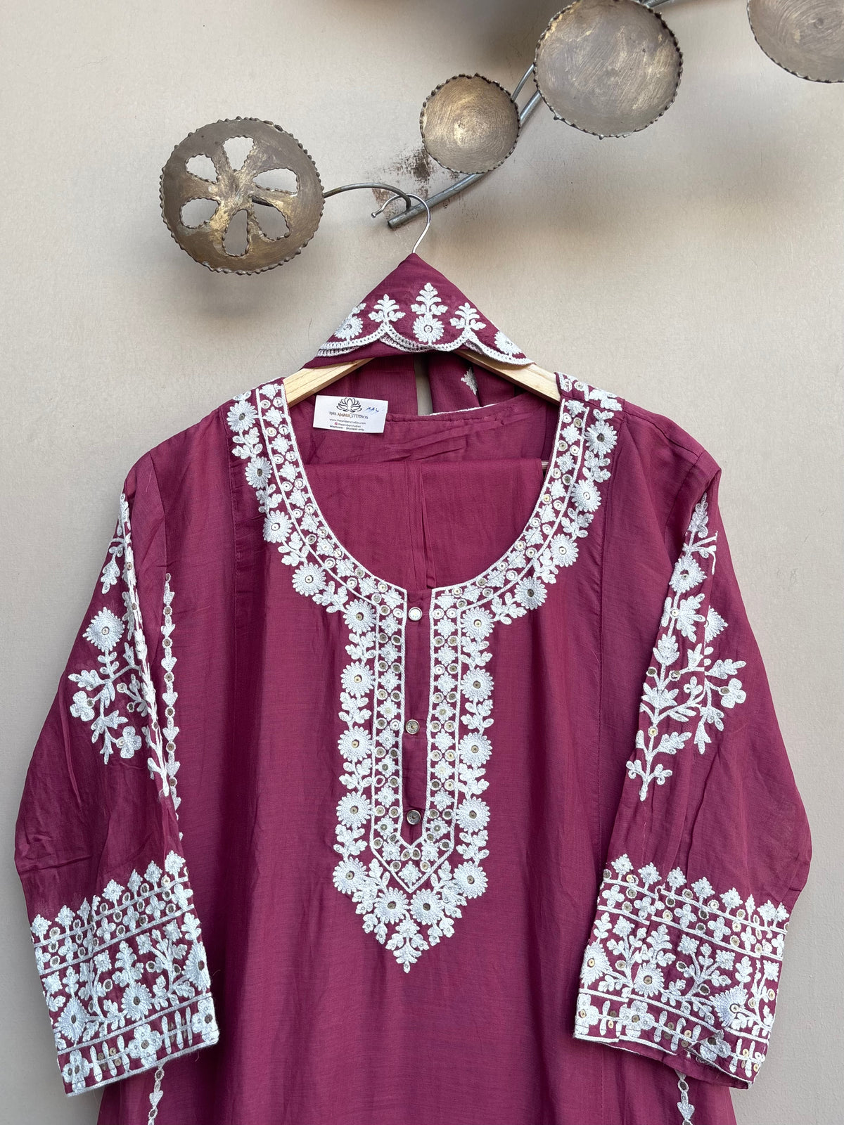A deep magenta-purple cotton kurta with intricate white floral embroidery on the neckline, sleeves, and cuffs, hanging on a wooden hanger against a textured beige wall.