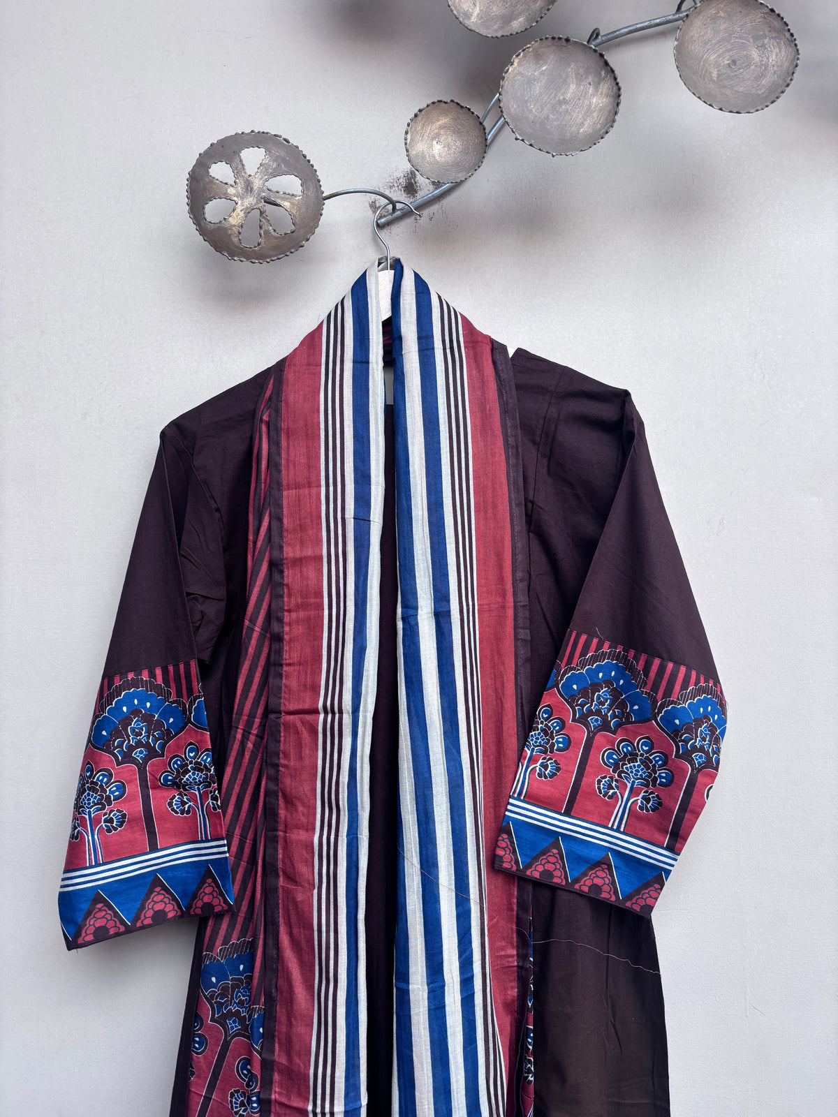 A dark brown traditional robe with a wide striped red, blue, and white shawl draped over the shoulders, featuring patterned red and blue sleeves, hanging on a wall under a decorative metal fixture.