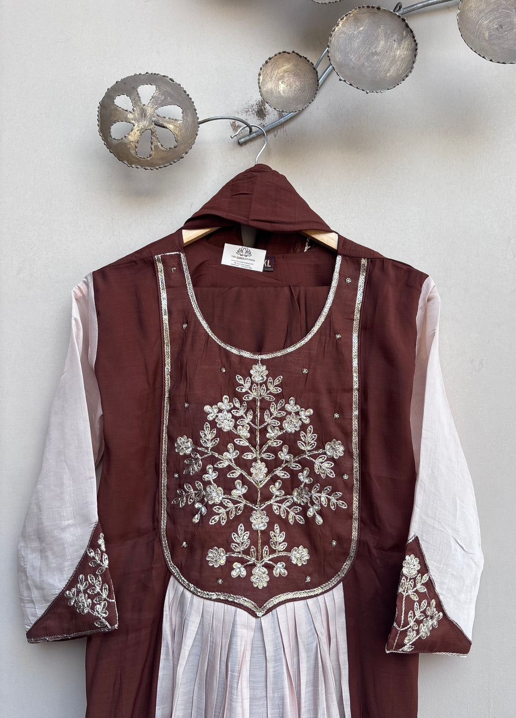 A brown and white embroidered tunic top with a hood, hanging on a wooden hanger against a light gray wall. The tunic features intricate silver floral embroidery on the chest and cuffs, with a white pleated skirt attached below.