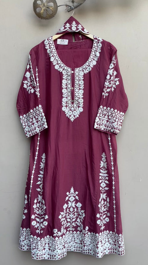 A maroon long kurta with intricate white embroidery, hanging on a wooden hanger against a plain beige wall.