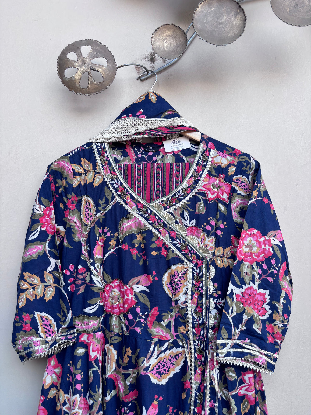 A navy blue floral and paisley wrap dress with pink, green, and gold patterns, featuring white lace trim, hanging on a decorative wall hook.