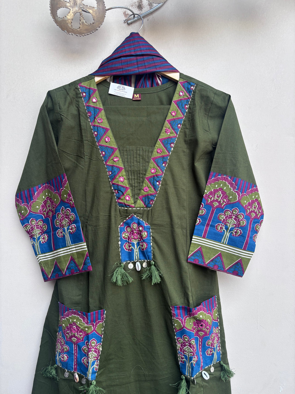 An olive green cotton tunic with vibrant blue, purple, and pink embroidered floral patterns on the neckline, cuffs, and pockets, hanging on a wooden hanger against a plain white wall.