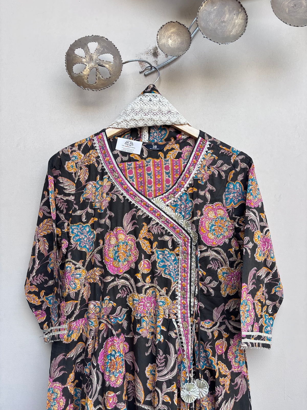 A black long-sleeve kurta with a vibrant multicolor floral print, hanging on a white lace-trimmed wooden hanger against a light gray wall.