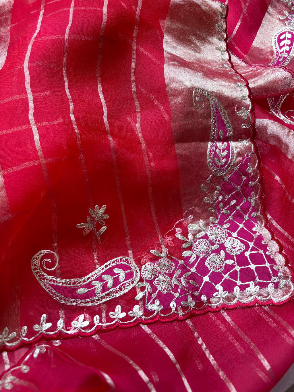 A close-up of a bright pink fabric with silver metallic stripes and ornate silver paisley and floral embroidery along a scalloped border.