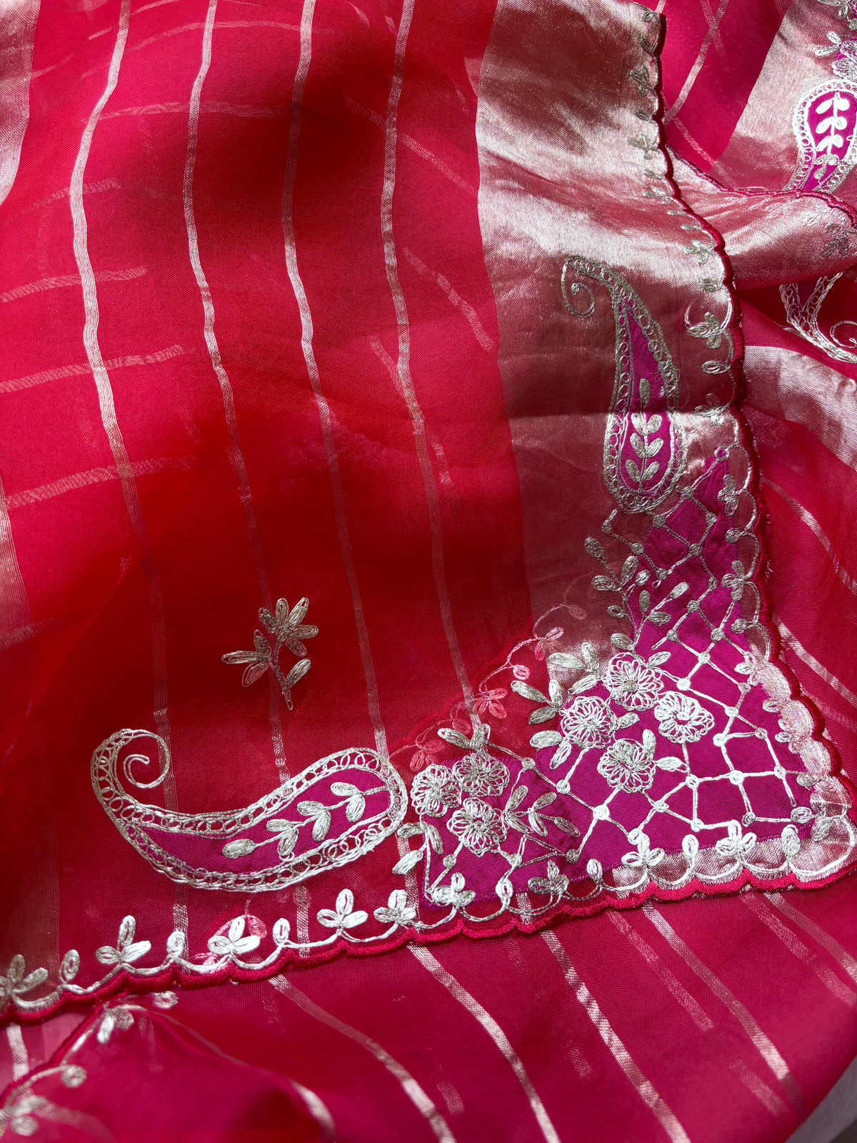 A close-up of a bright pink fabric with silver metallic stripes and ornate silver paisley and floral embroidery along a scalloped border.