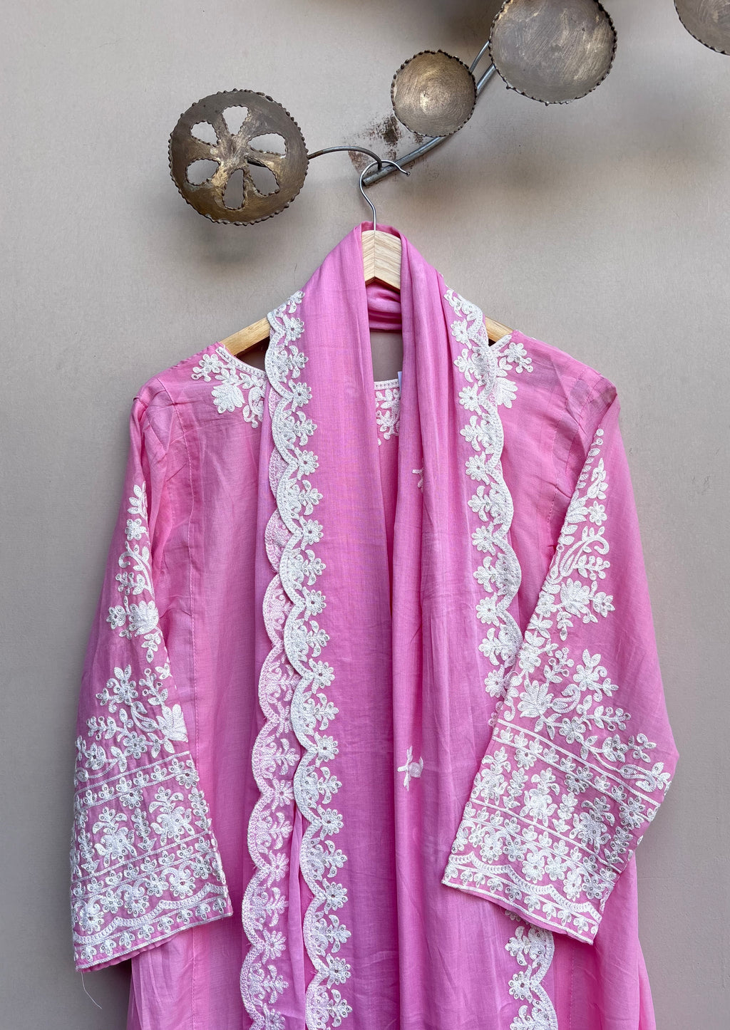 A pink traditional embroidered garment with a matching scarf draped over a hanger, featuring intricate white floral embroidery on the sleeves, collar, and front border, set against a neutral wall with decorative metal wall art.