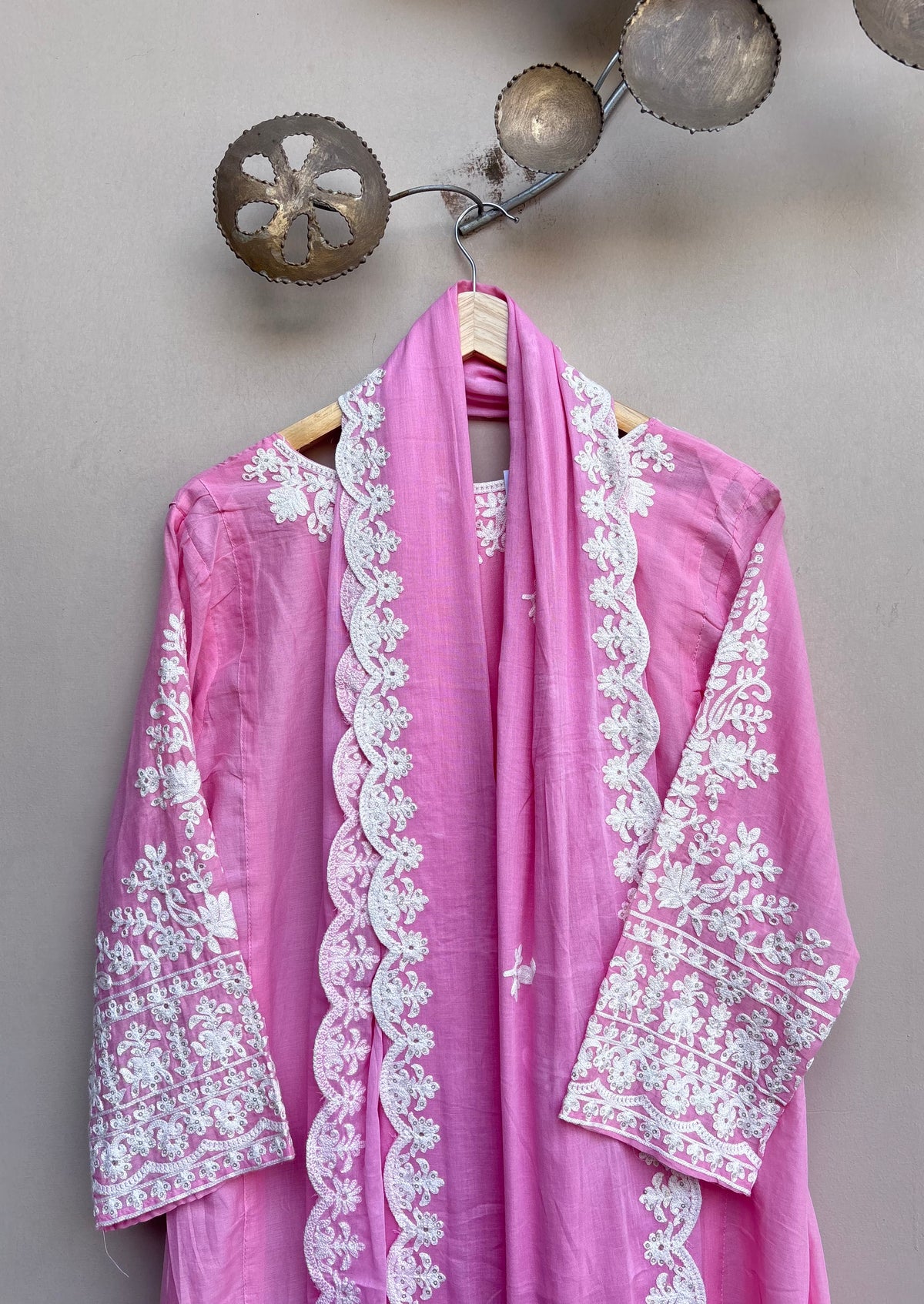 A pink traditional embroidered garment with a matching scarf draped over a hanger, featuring intricate white floral embroidery on the sleeves, collar, and front border, set against a neutral wall with decorative metal wall art.