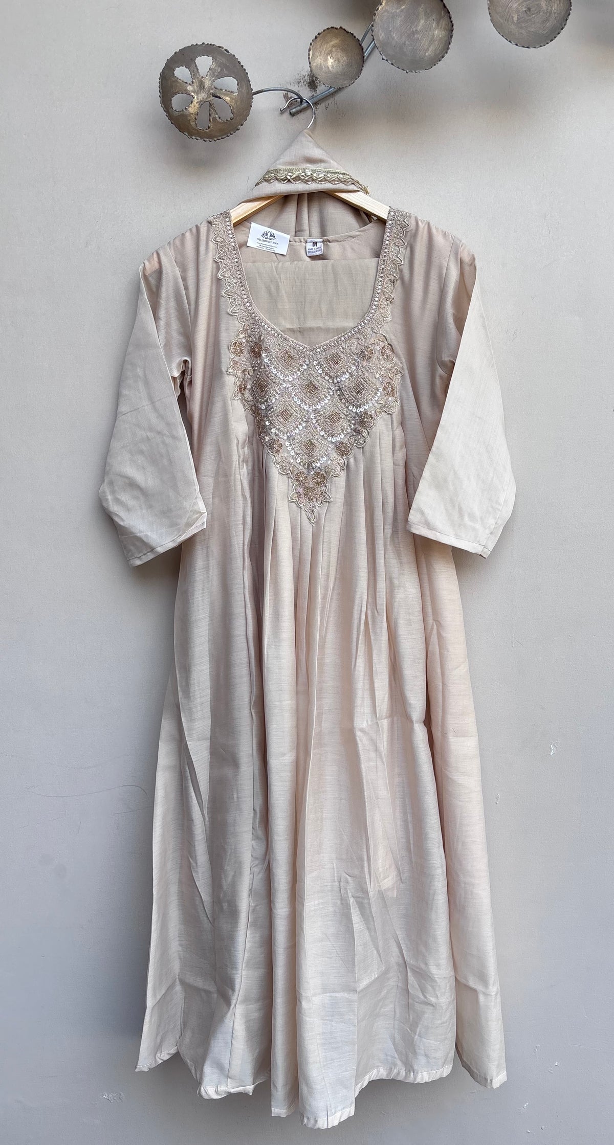 A light beige linen tunic dress with embroidered detailing on the chest, hanging on a wooden hanger against a textured gray wall.