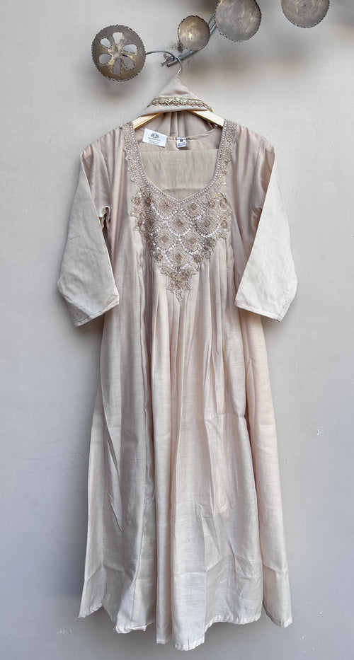 A light beige linen tunic dress with embroidered detailing on the chest, hanging on a wooden hanger against a textured gray wall.