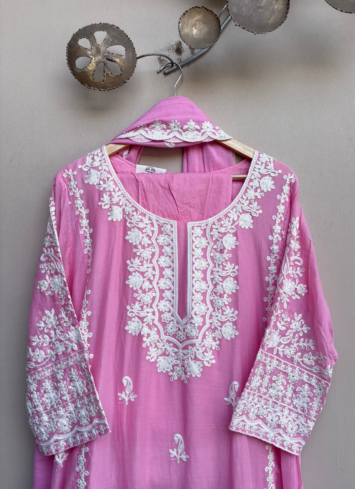 A pink traditional long kurta with intricate white floral embroidery, hanging on a wooden hanger against a neutral gray wall.