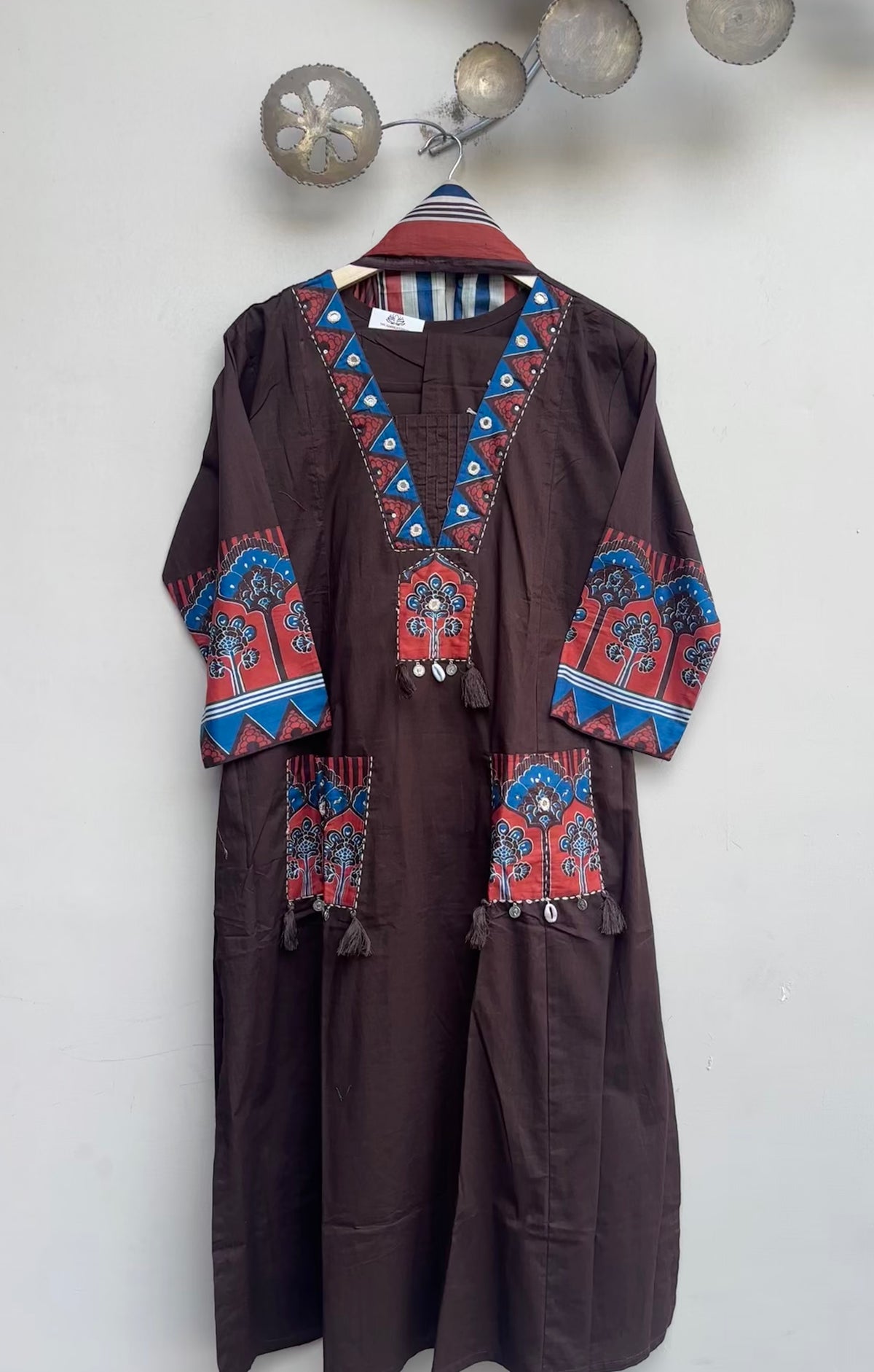 A dark brown traditional long robe with colorful red, blue, and white embroidered patterns on the collar, cuffs, and pockets, hanging on a wall.