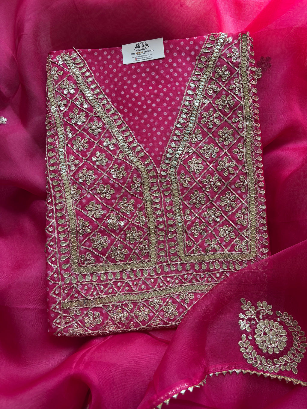 A folded bright pink embroidered fabric garment, likely a kurta or dress panel, with extensive metallic gold zari embroidery and mirror work, displayed on a matching pink satin fabric background.