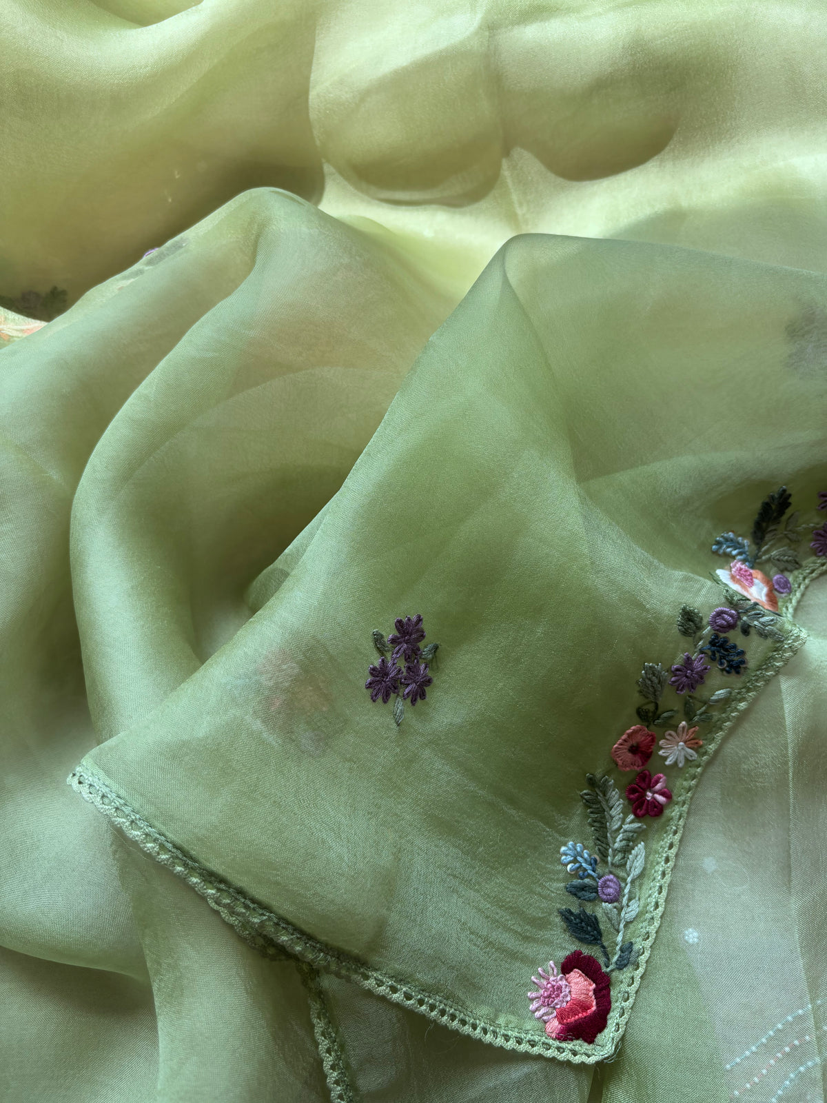 A close-up of a pale yellow-green sheer fabric, likely organza or tulle, with embroidered floral details in purple, pink, and green along one edge.