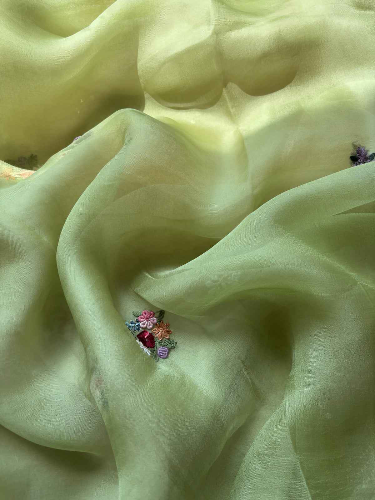 A close-up of draped pale yellow-green sheer fabric with a small colorful embroidered floral detail near the lower center.