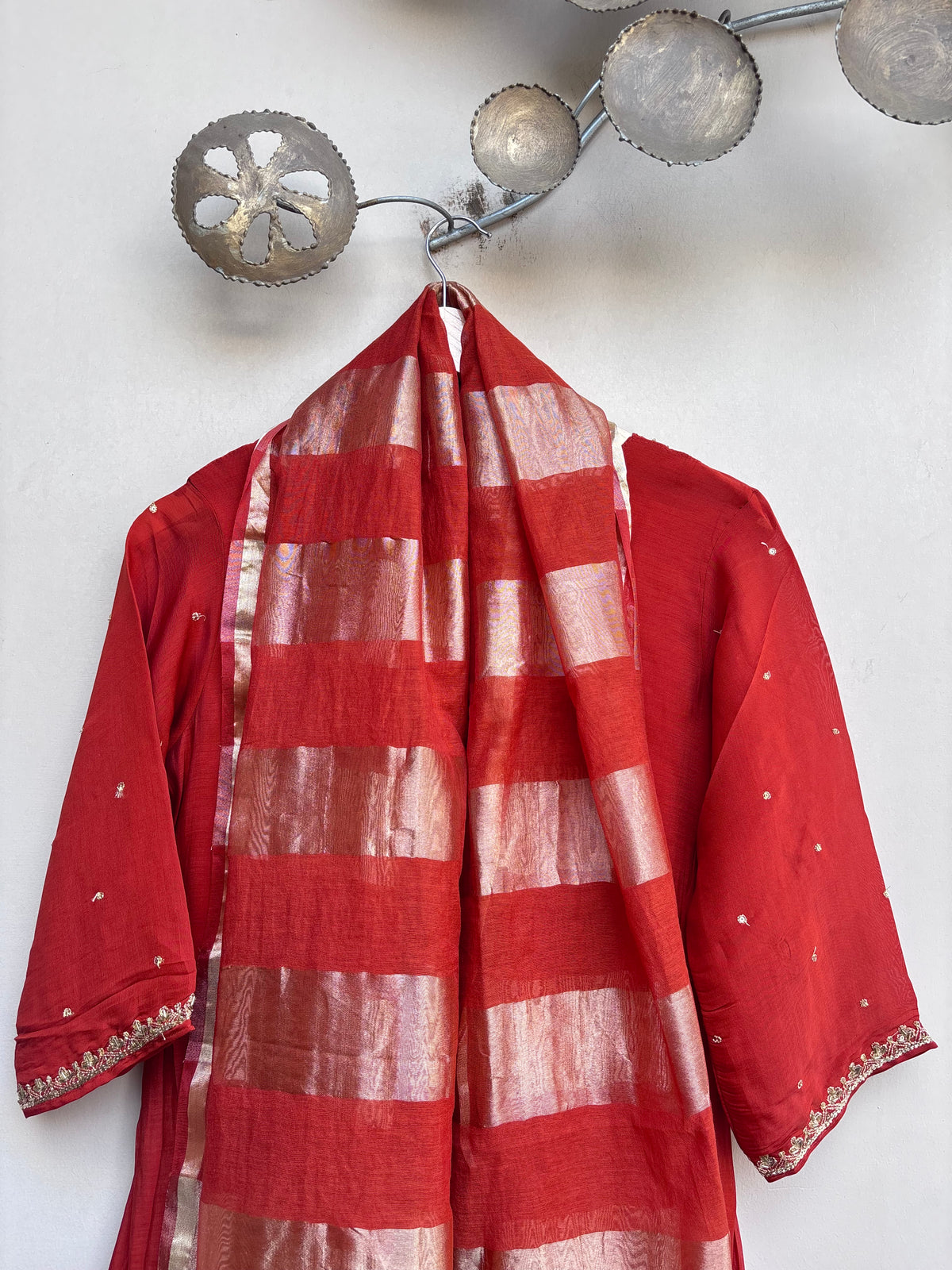 A red traditional garment with a matching red and gold striped dupatta draped over a hanger against a white wall.