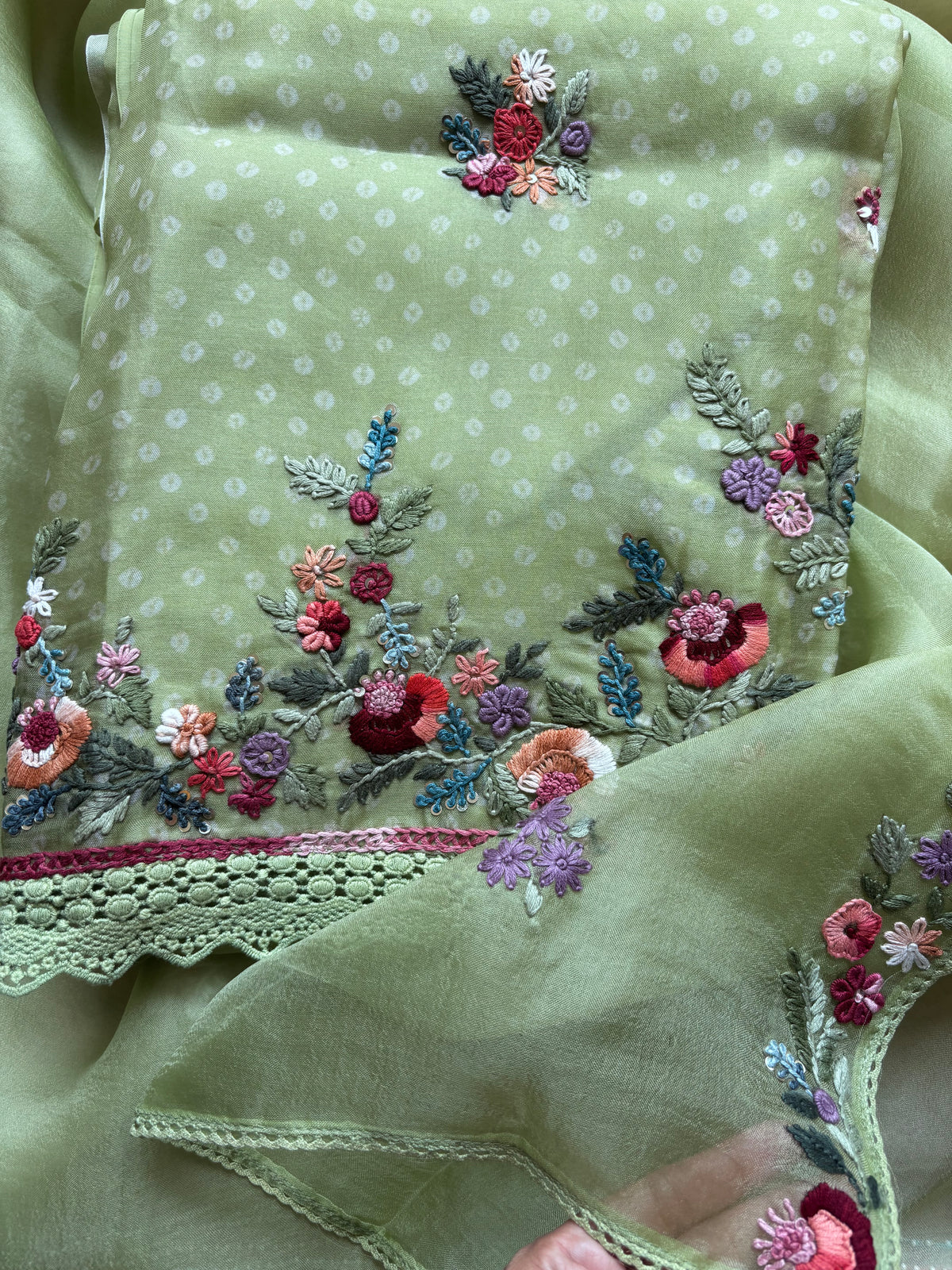 A close-up of a light sage green embroidered fabric, likely part of a traditional ethnic outfit set. The fabric features a subtle white polka dot pattern, a scalloped lace border, and vibrant floral embroidery in shades of pink, red, purple, and green.