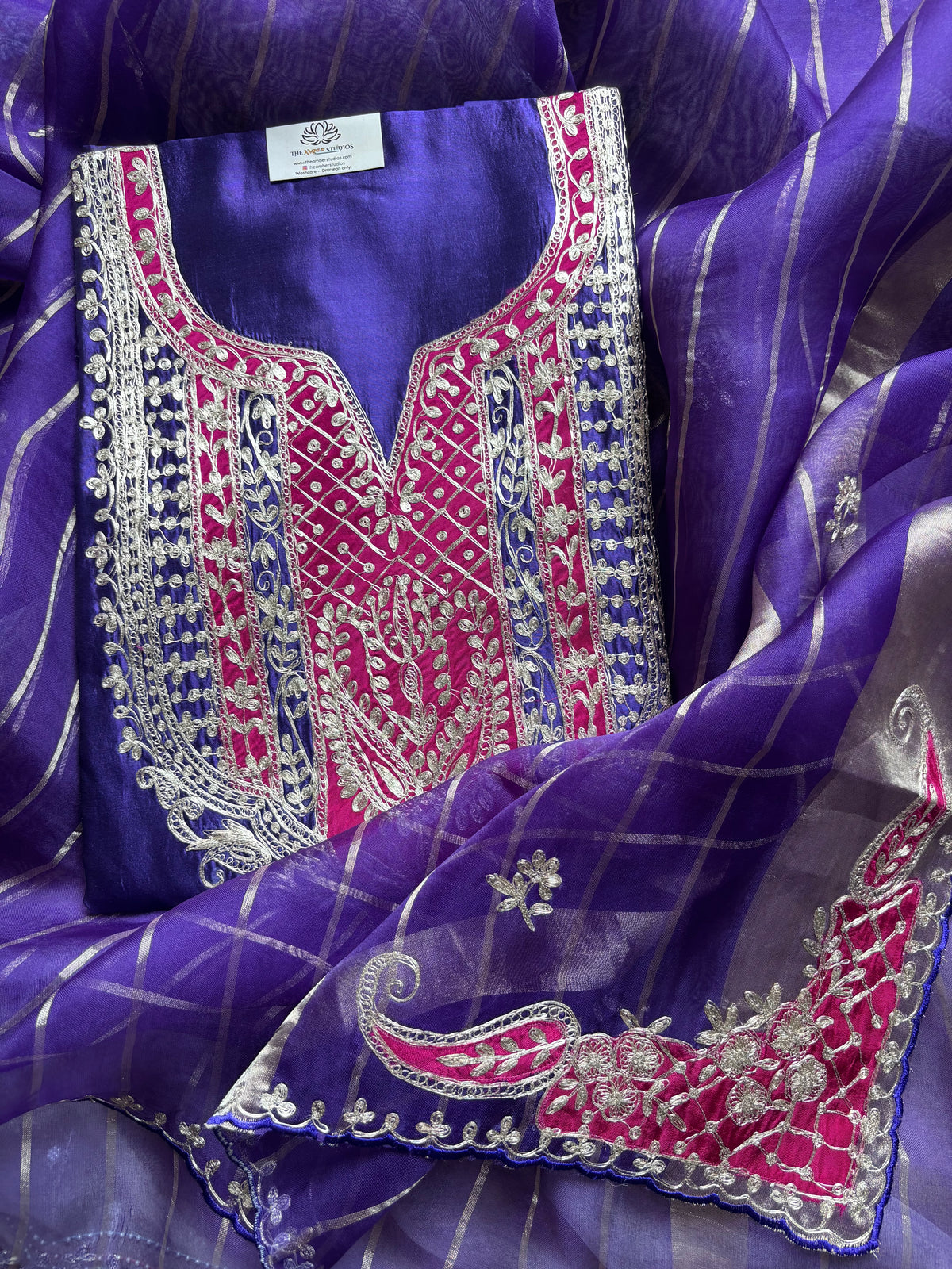 A purple traditional embroidered garment laid flat on a matching sheer purple dupatta. The fabric features prominent pink and silver mirror embroidery with intricate geometric and floral patterns around the neckline and borders.