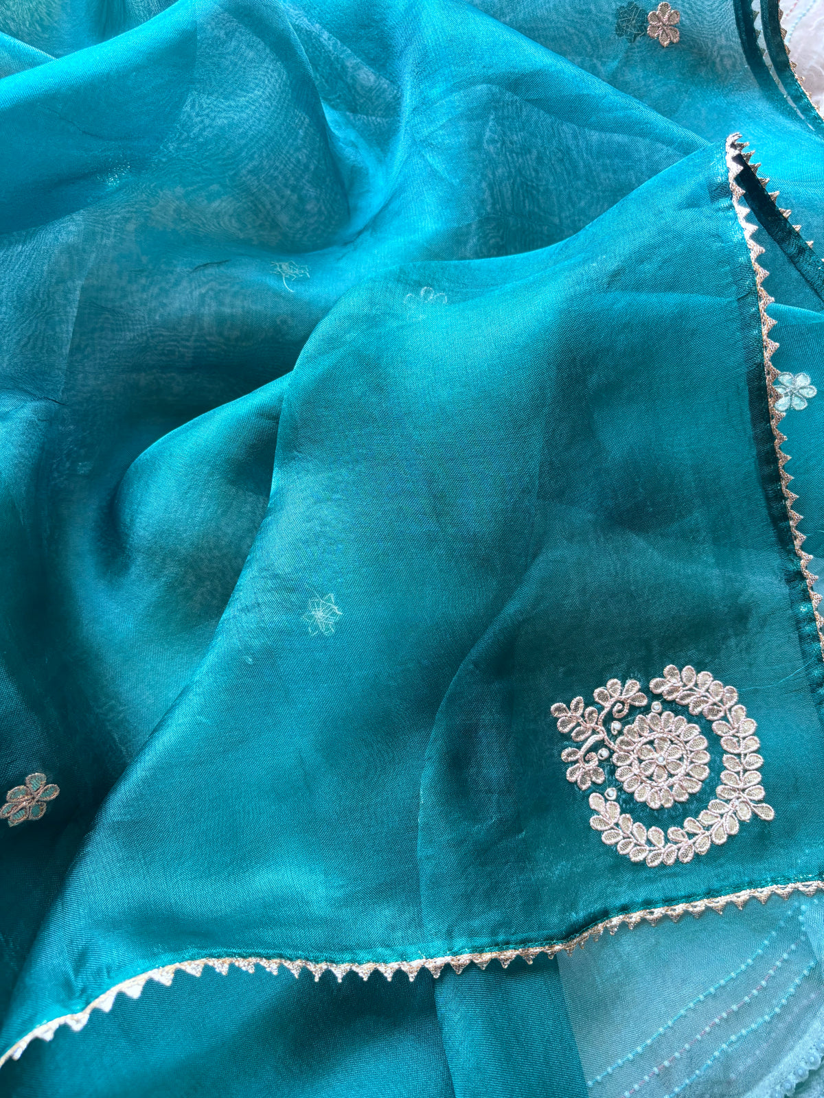 A close-up of a teal blue sheer fabric, likely organza or georgette, featuring delicate silver floral embroidery and a scalloped embroidered border.