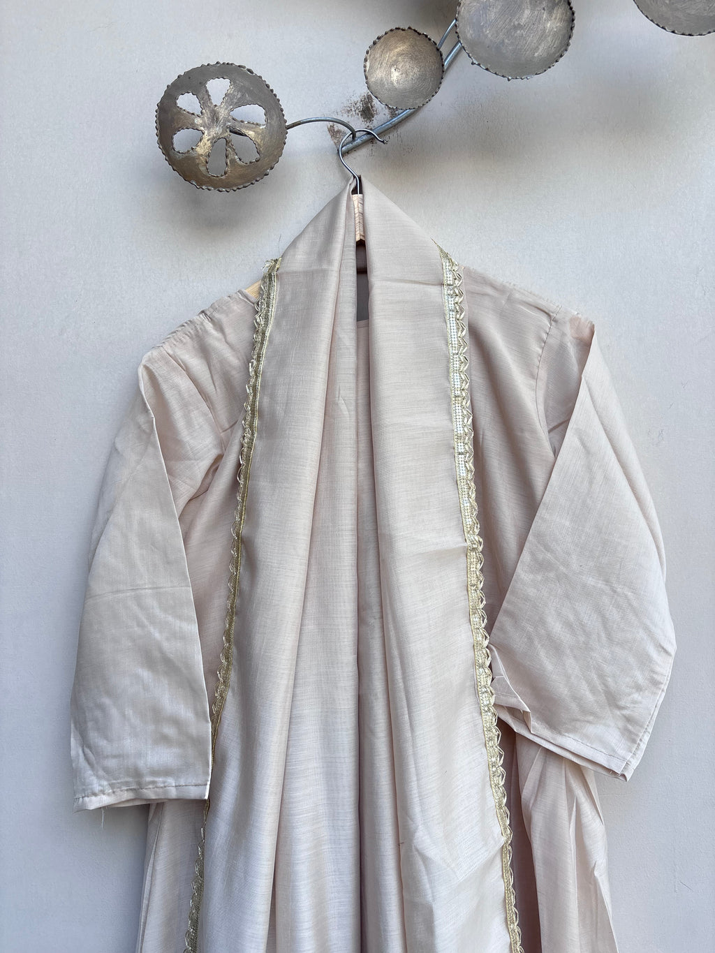 A cream-colored linen kimono robe with gold embroidered trim hanging on a decorative metal wall hook against a textured off-white wall.