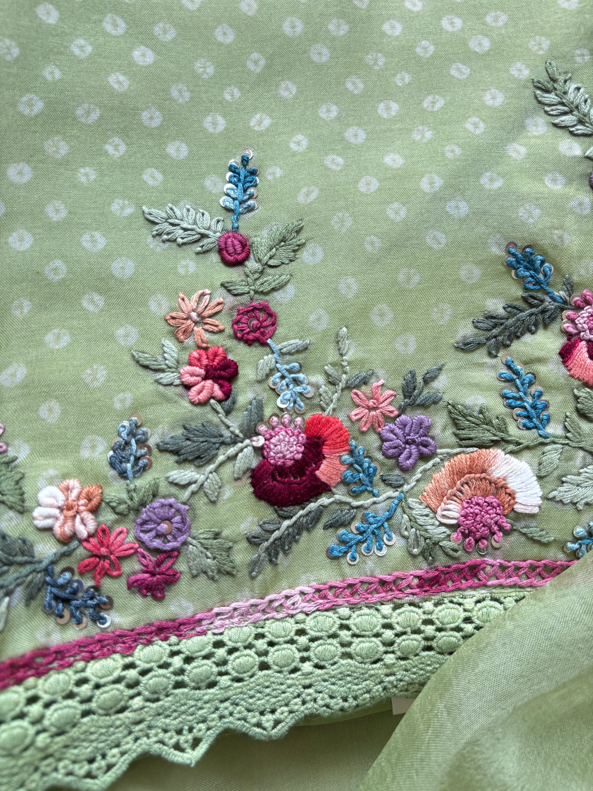 Close-up of a light green embroidered fabric with a floral pattern, featuring pink, red, blue, and purple embroidery, a pink lace border, and a matching sheer green fabric in the corner.