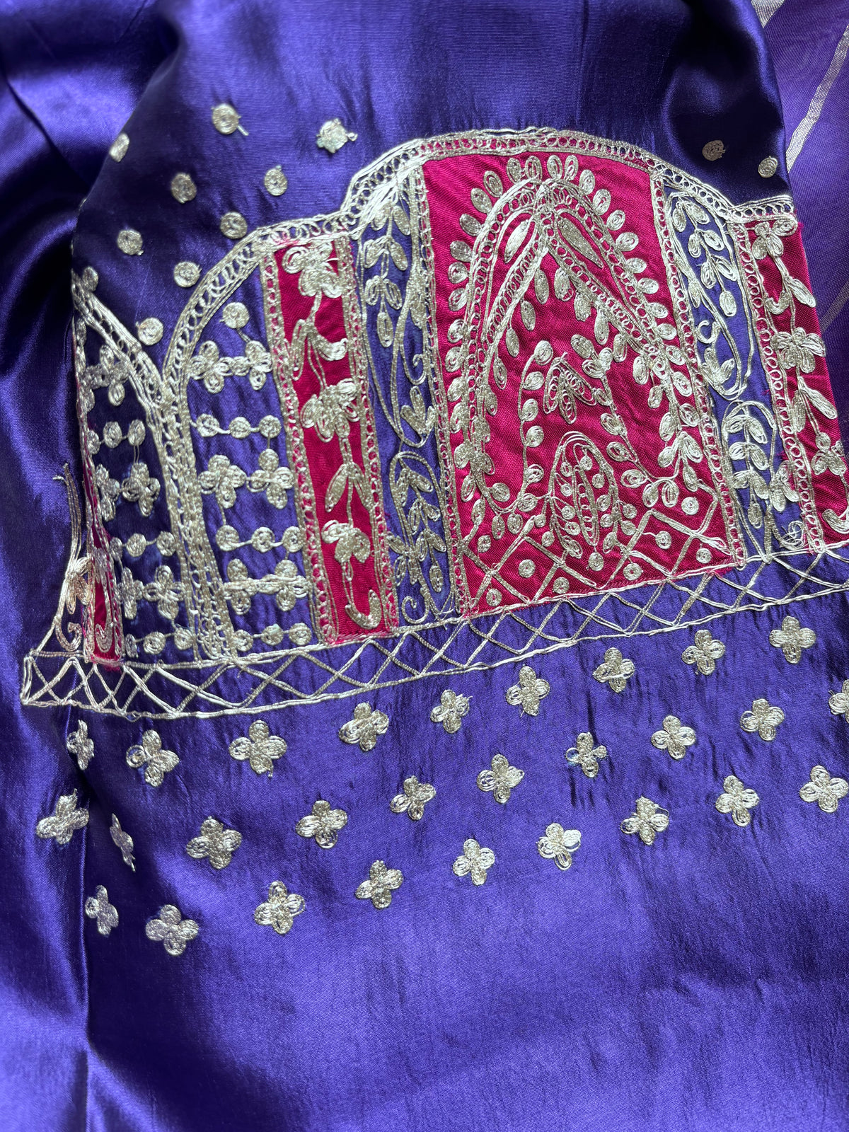 A close-up of a purple satin fabric garment featuring intricate silver embroidery and a central red and blue embroidered panel.