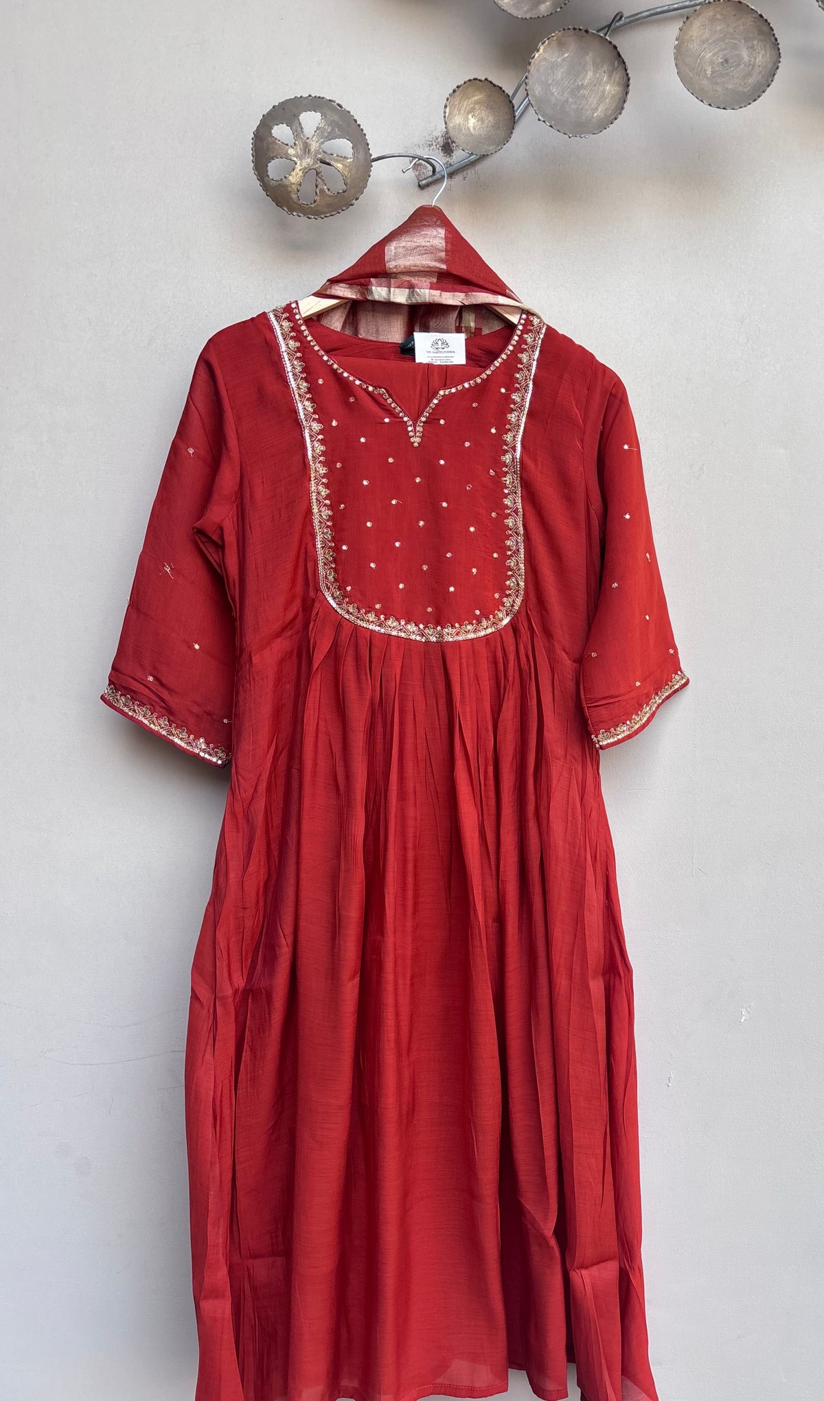 A red cotton kurta dress with gold embroidery and mirror work on the chest, hanging on a hanger against a light gray wall with decorative metal spheres.