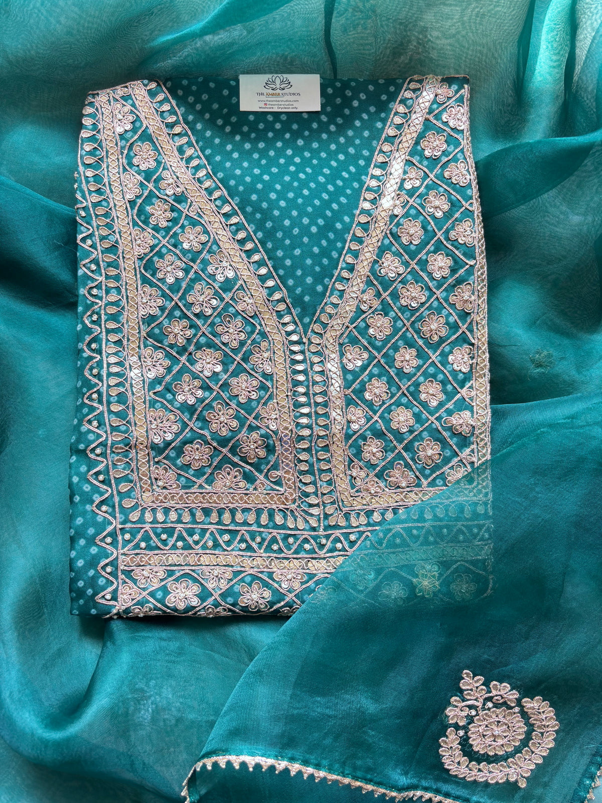 A folded teal blue and white printed ethnic garment with ornate silver and white embroidery, displayed on a matching teal fabric background.