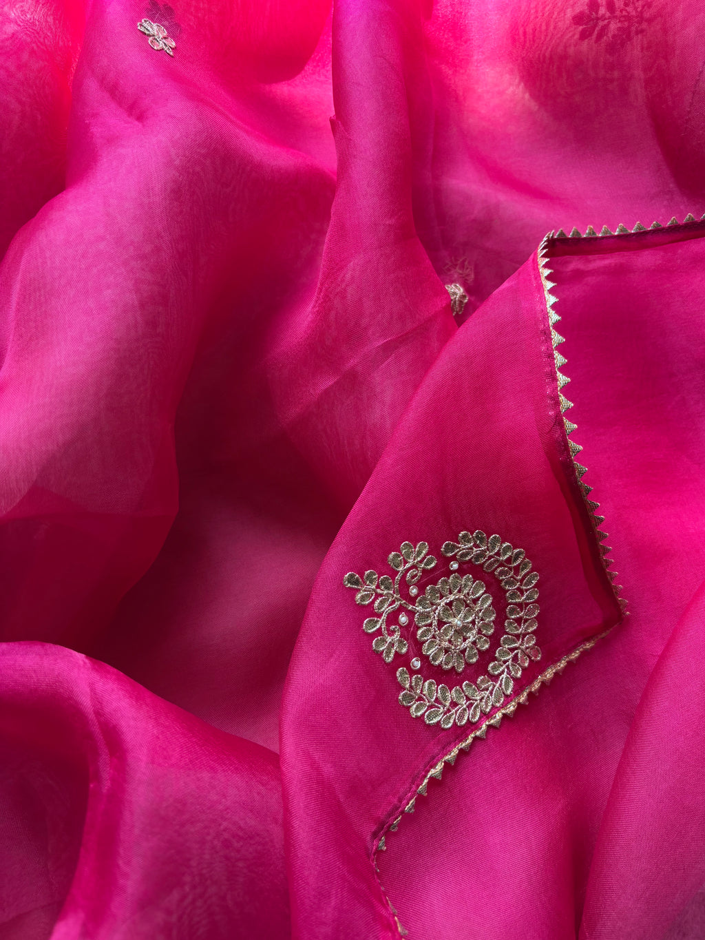 A close-up of bright pink fabric with a matching embroidered section featuring metallic gold floral embroidery and a zigzag border.
