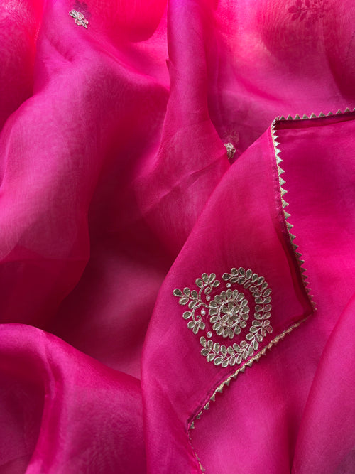 A close-up of bright pink fabric with a matching embroidered section featuring metallic gold floral embroidery and a zigzag border.