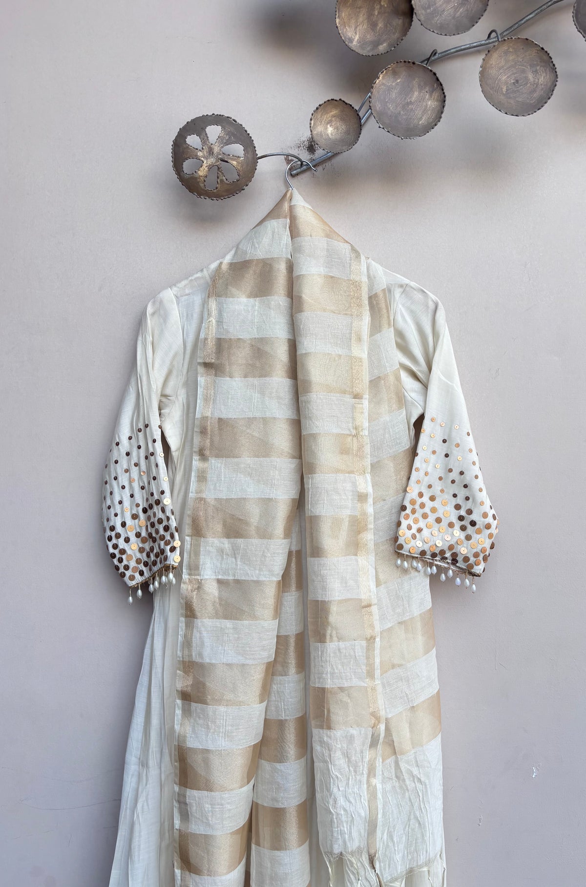 A cream and beige striped shawl draped over a white kurta with gold polka dot sleeve embellishments, hanging on a decorative branch against a light gray wall.