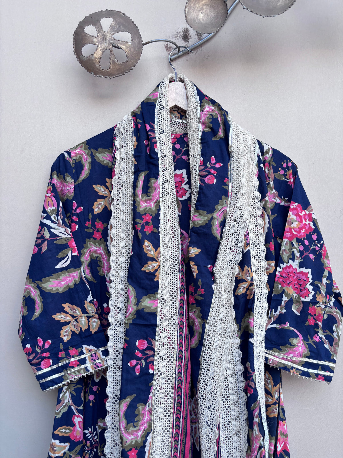 A navy blue floral kimono robe with pink and green paisley patterns, featuring white lace trim down the front, hanging on a white hanger against a light gray wall.