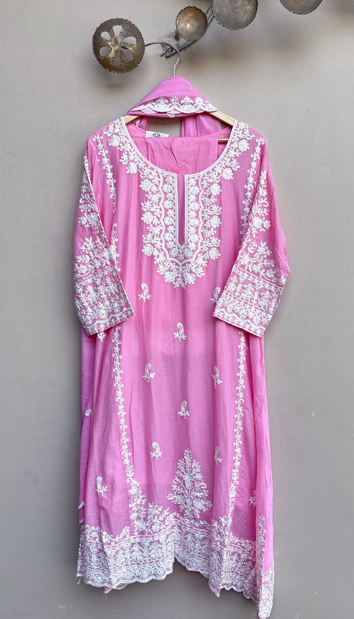 A pink traditional kurta with intricate white floral embroidery, hanging on a wooden hanger against a textured gray wall.