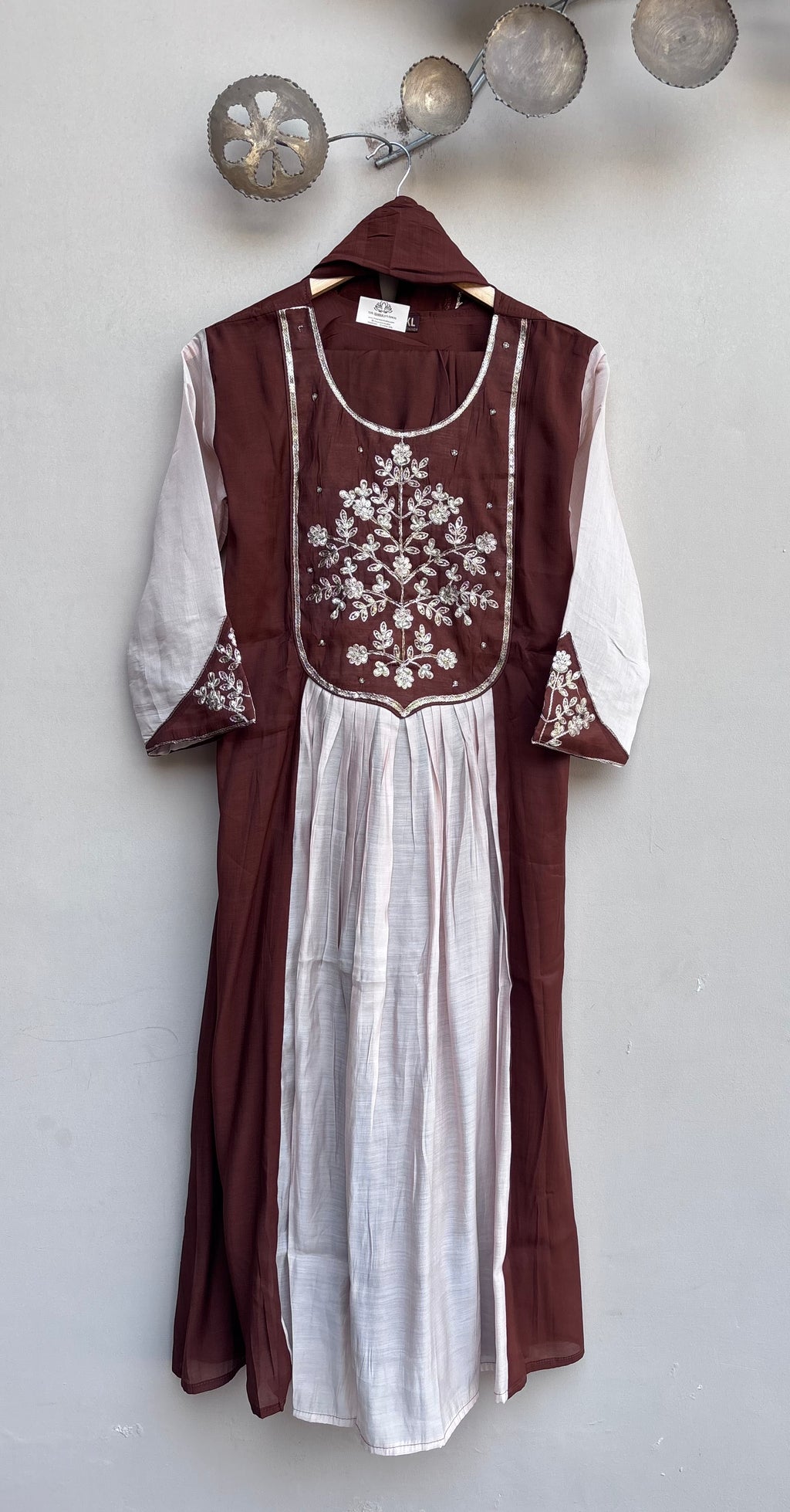 A traditional long dress with a deep brown outer layer and a white inner dress, featuring white floral embroidery on the chest and sleeve cuffs, hanging against a light gray wall.