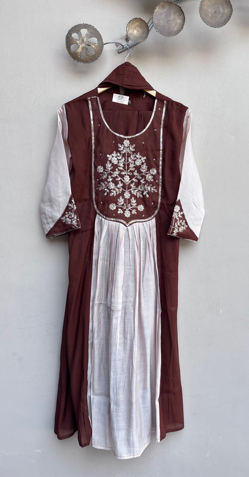 A traditional long dress with a deep brown outer layer and a white inner dress, featuring white floral embroidery on the chest and sleeve cuffs, hanging against a light gray wall.