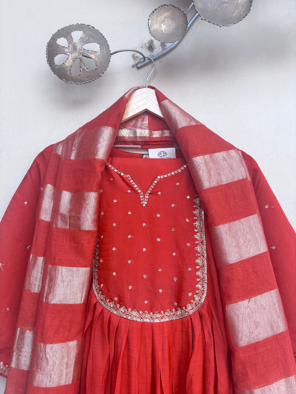 A red traditional outfit with a matching striped red and white dupatta draped over a hanger, set against a light gray wall with decorative metal wall art.