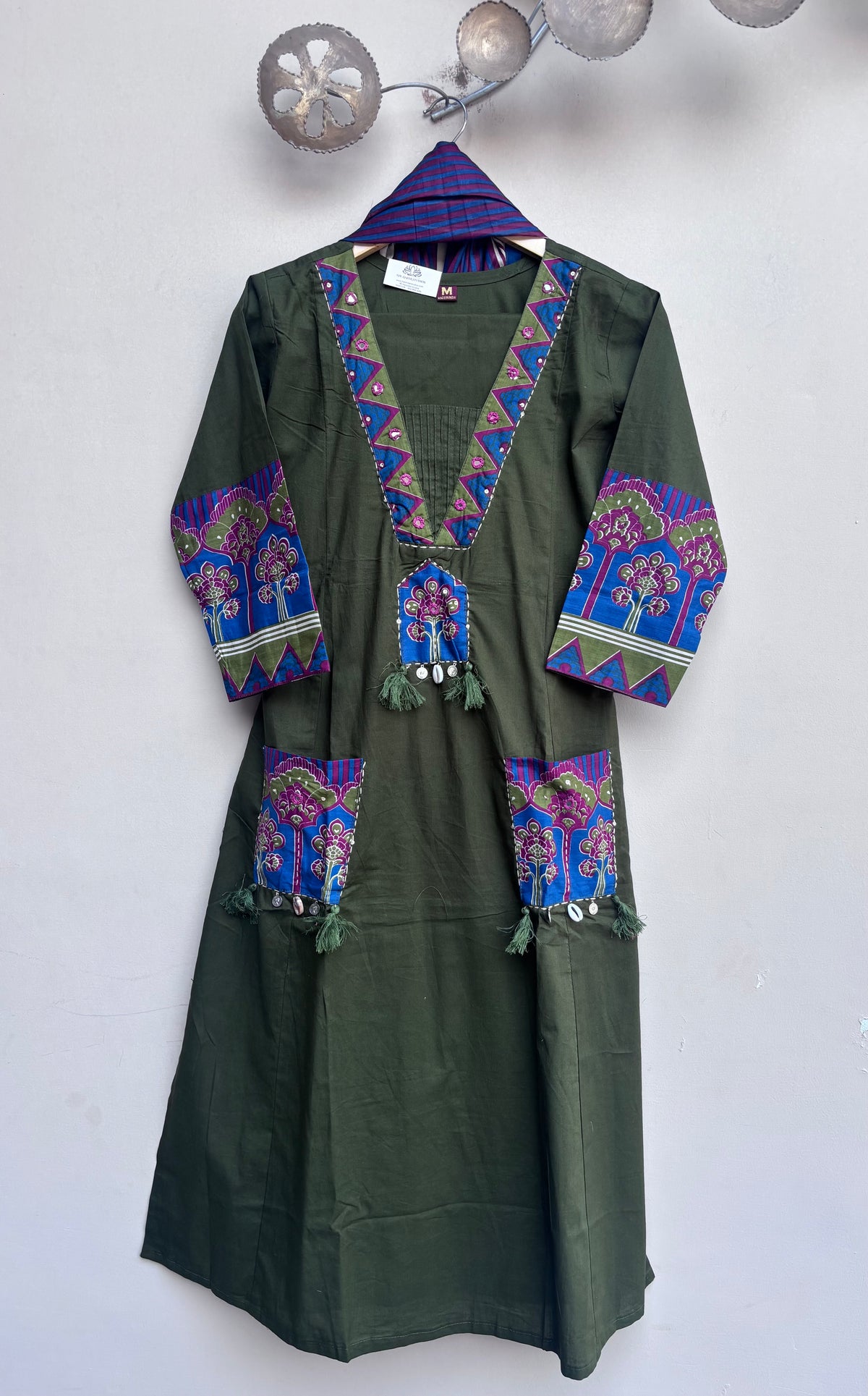 An olive green long-sleeve tunic or kaftan dress with colorful embroidered details on the neckline, cuffs, and pockets, hanging on a patterned hanger against a plain wall.