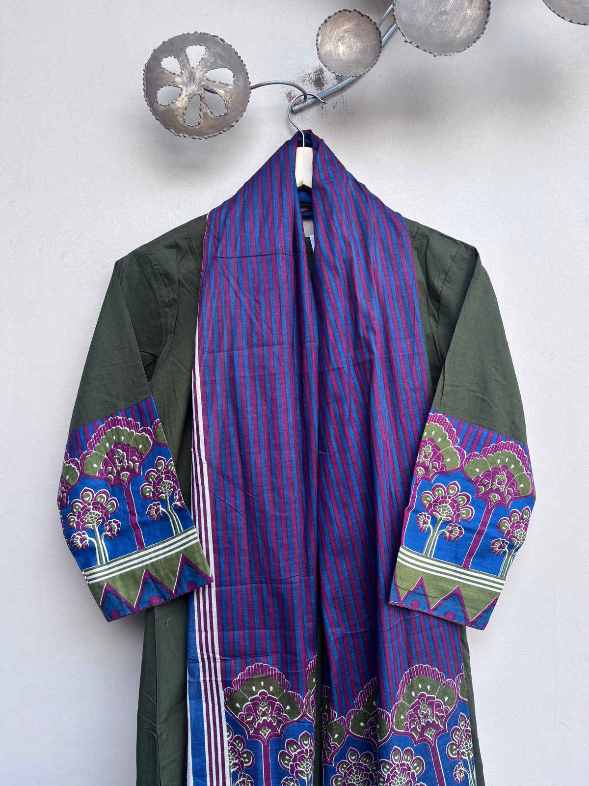 A traditional open-front robe with a purple and blue vertically striped center panel, olive green sleeves, and colorful floral mandala patterns on the cuffs and hem, hanging on a decorative metal wall hook against a white wall.