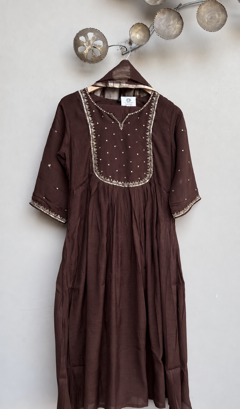 A dark brown cotton kurta dress with silver mirror work embroidery on the yoke and sleeve cuffs, hanging on a wooden hanger against a light gray wall.