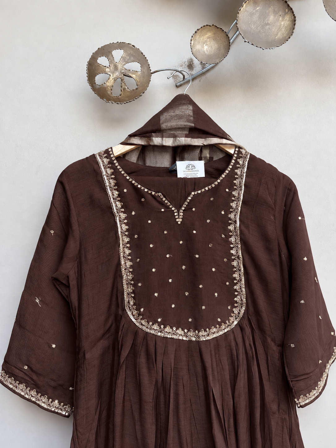 A dark brown cotton tunic dress with gold embroidery and mirror work on the chest, hanging on a wooden hanger against a textured off-white wall with decorative metal wall art.