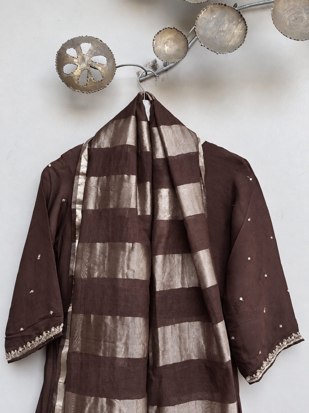 A brown and metallic silver striped traditional tunic with matching draped scarf, displayed on a hanger against a light wall with decorative metal wall art.