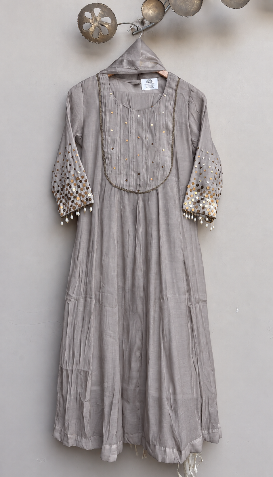 A light gray linen kurta with gold and silver mirror work embroidery on the yoke and sleeves, hanging on a wooden hanger against a neutral gray wall.