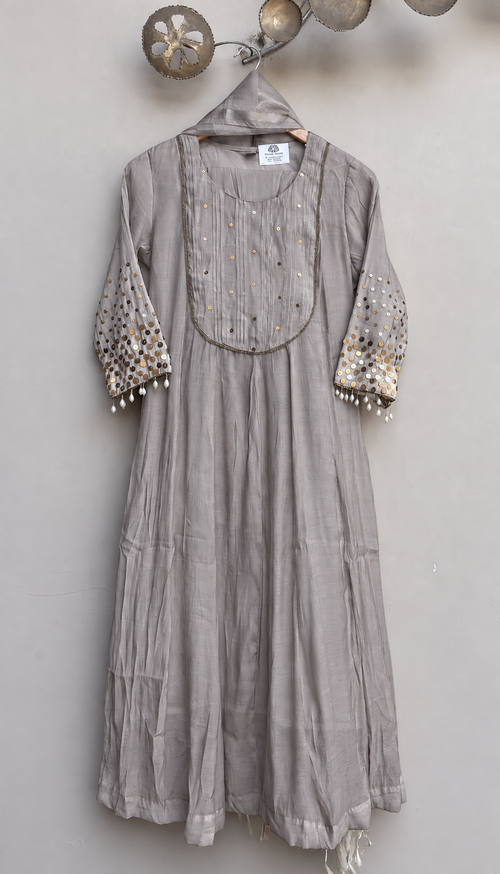 A light gray linen kurta with gold and silver mirror work embroidery on the yoke and sleeves, hanging on a wooden hanger against a neutral gray wall.
