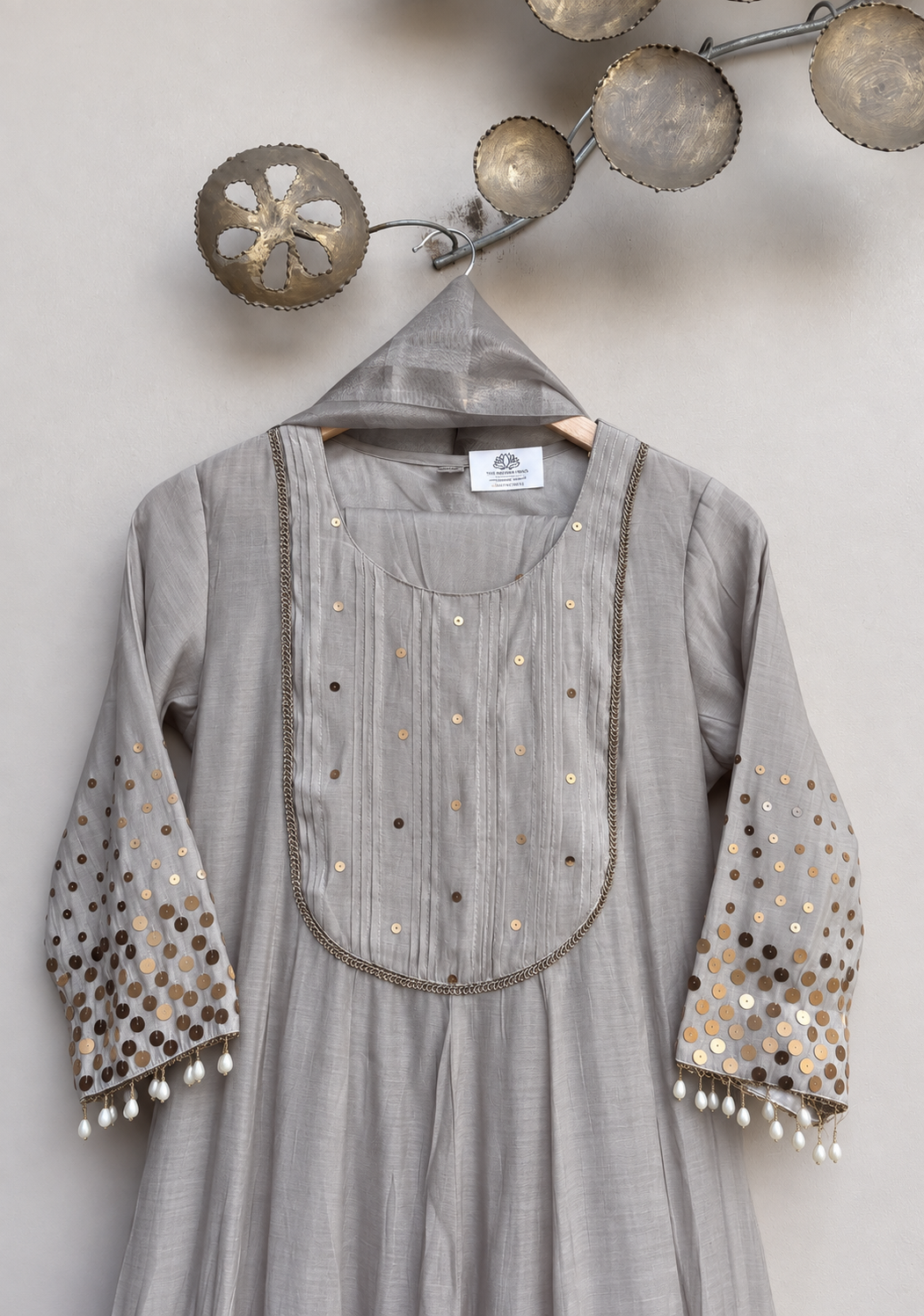 A light gray three-quarter sleeve kurta with gold and silver sequin embellishments, hanging on a wooden hanger against a neutral gray wall with decorative wall art.