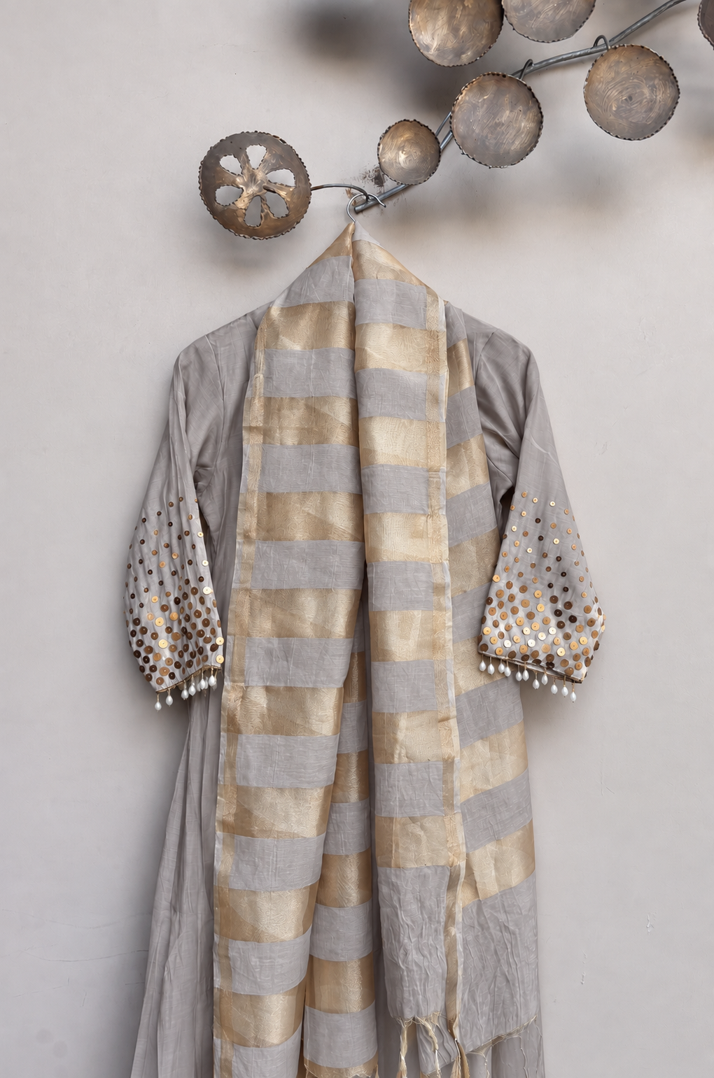 A light gray three-piece traditional outfit with a matching striped scarf, featuring gold-embroidered and pearl-embellished sleeves, displayed against a neutral wall with decorative metal wall art.