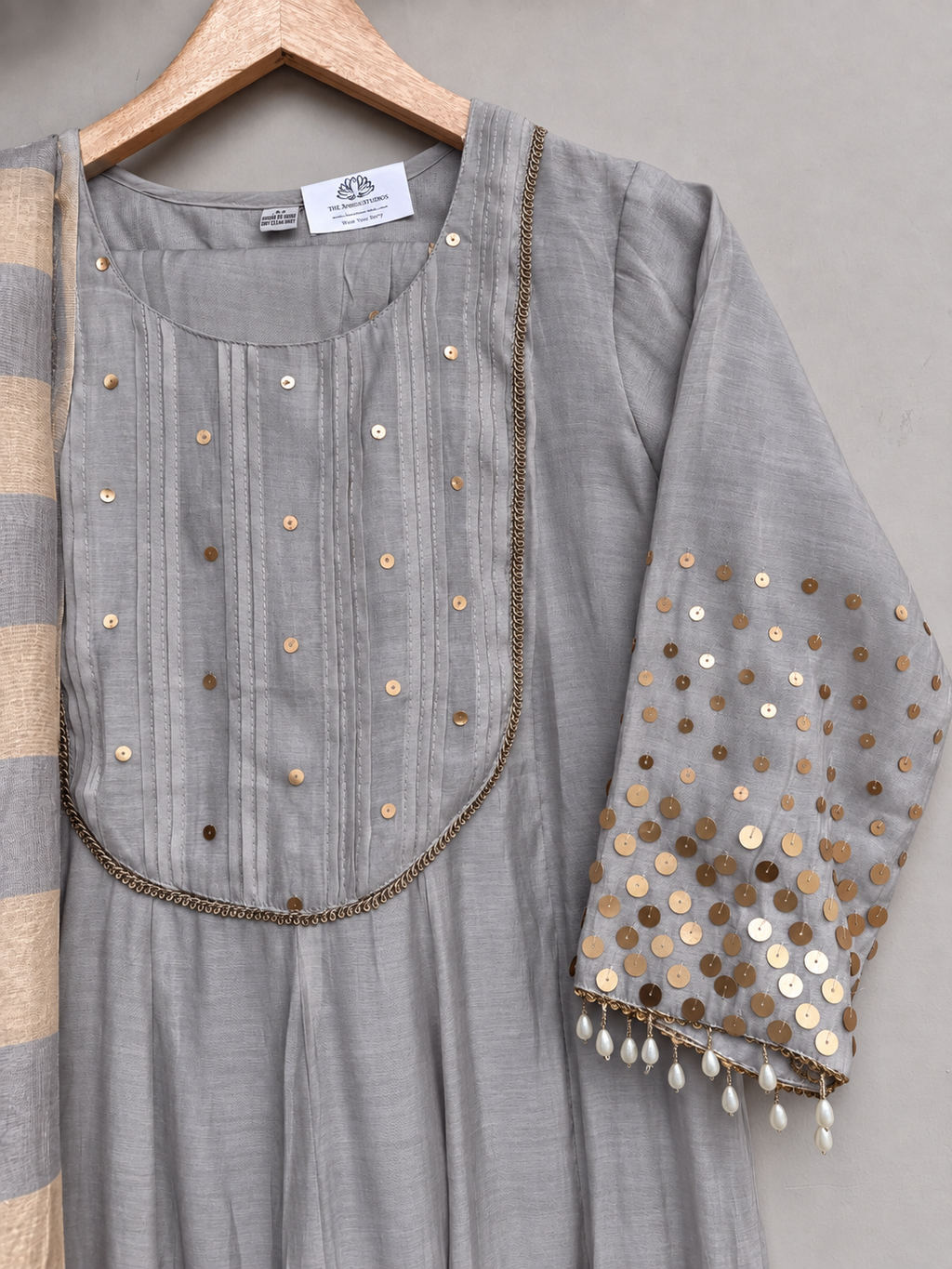 A grey kurta with metallic sequin embellishments on the yoke and sleeve, hanging on a wooden hanger against a neutral gray wall.