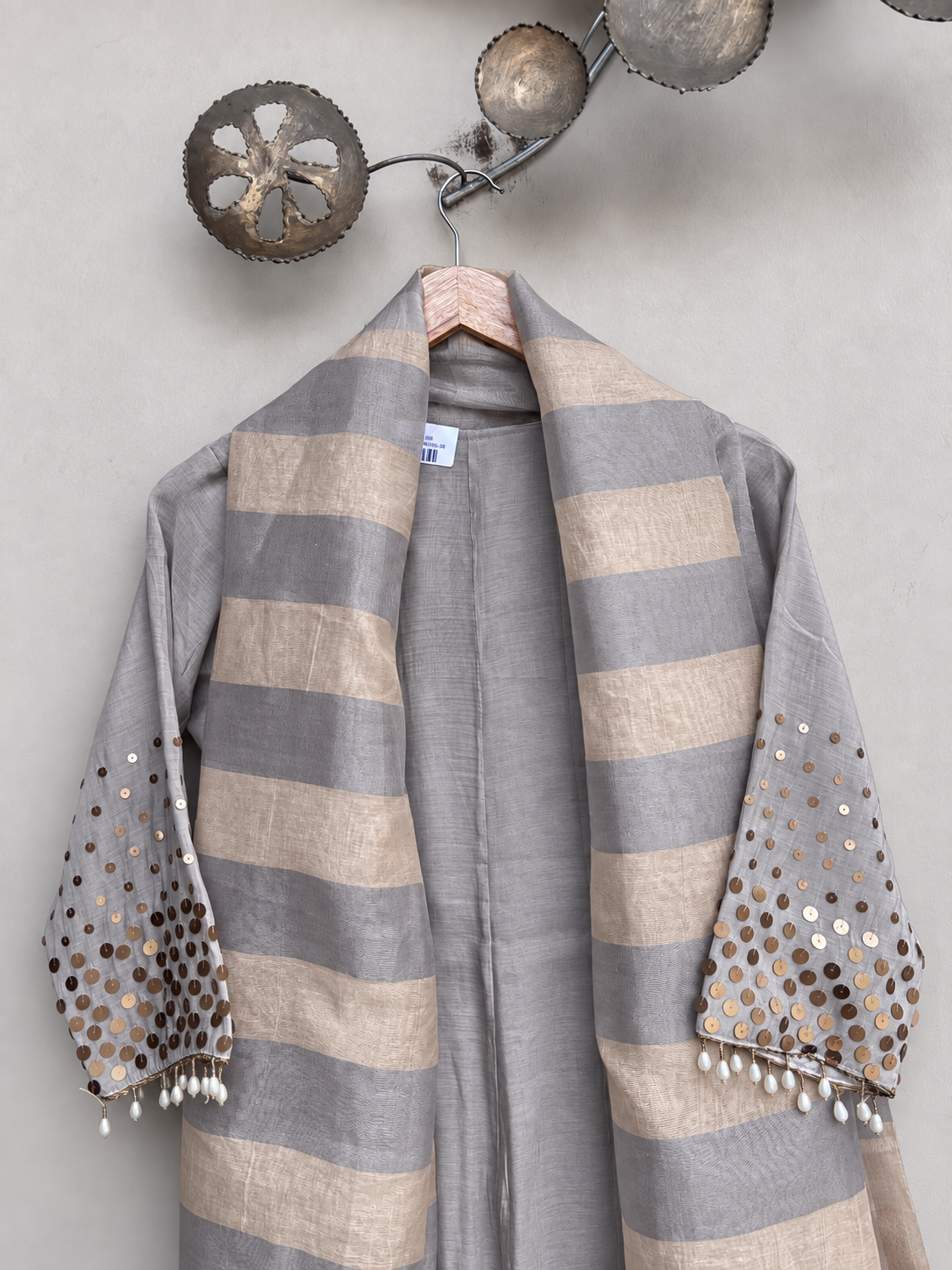 A striped grey shawl with sequined and tassel details, hanging on a wooden hanger against a textured light gray wall.