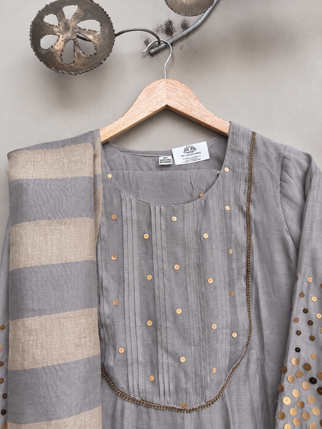A grey traditional Indian outfit with a striped dupatta, hanging on a wooden hanger against a textured wall.