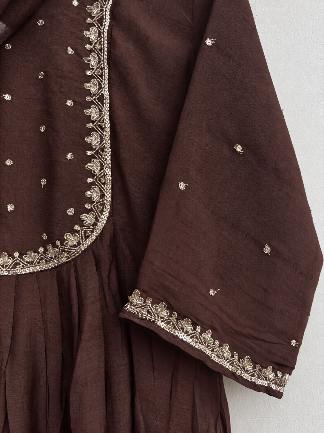A close-up of a brown traditional garment, likely a kurta or anarkali suit, featuring gold and silver embroidery along the border and scattered small metallic motifs on the fabric.