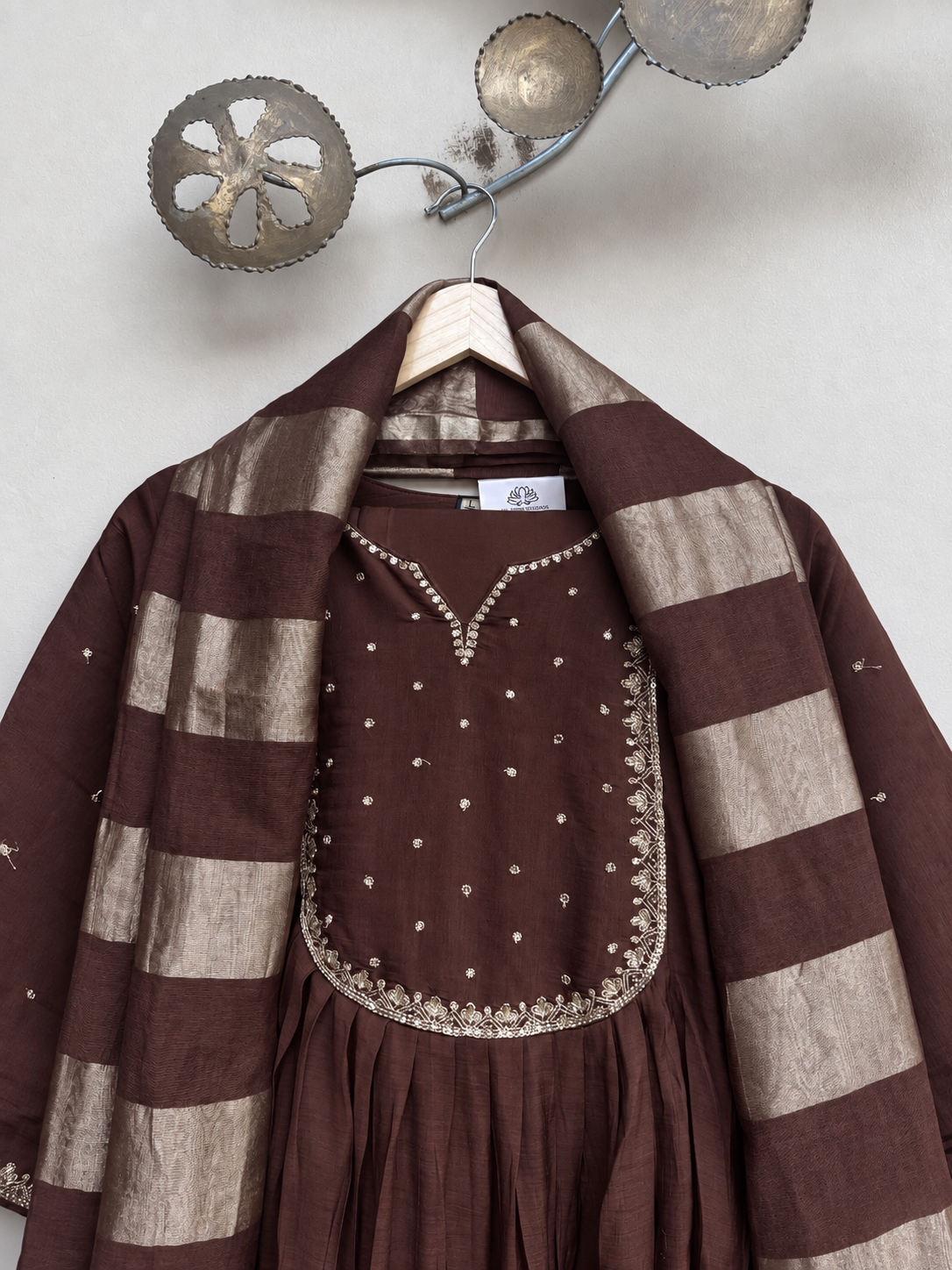 A brown and beige striped shawl draped over a brown embroidered kurta on a wooden hanger against a light gray wall.
